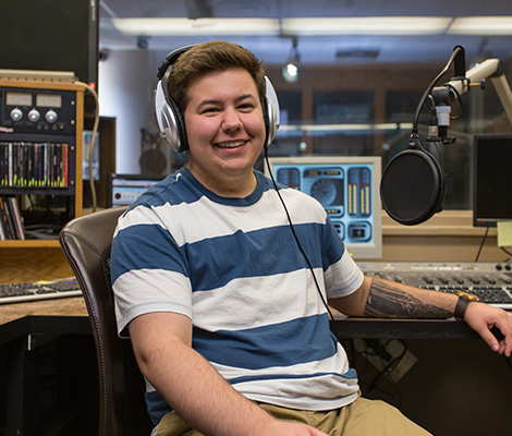 A male student wearing headphoness sits in front of a microphones in a recording studio.